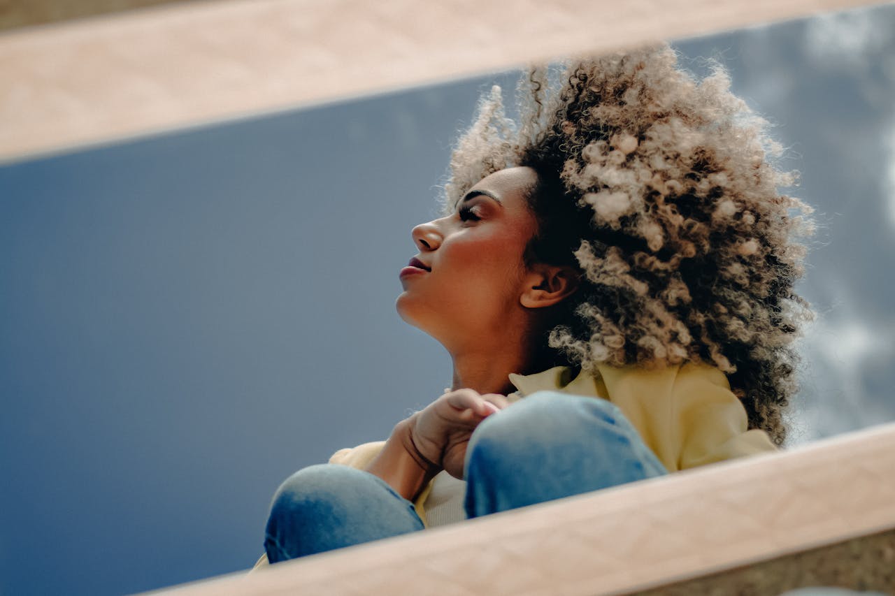 A woman with curly hair gazes upwards in a reflective mirror shot against a sky backdrop.