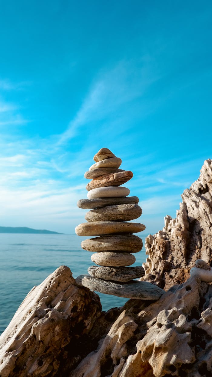 A stack of balanced stones on a rocky seashore with a clear blue sky and sea in the background.