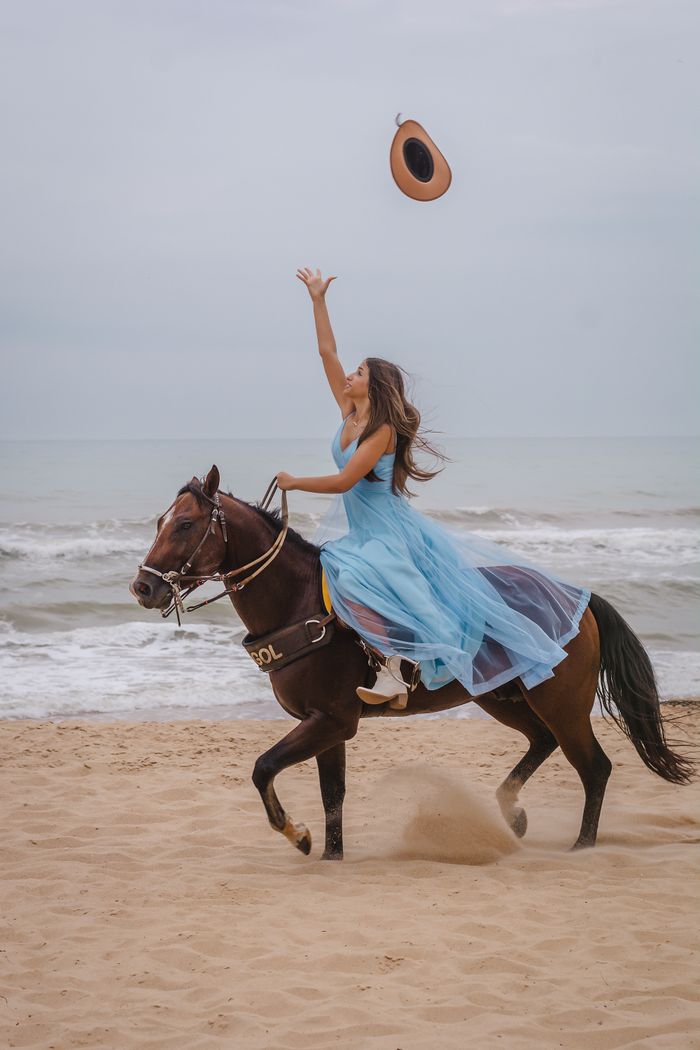 Woman in a blue dress riding a horse on a sandy beach, throwing a hat in the air.